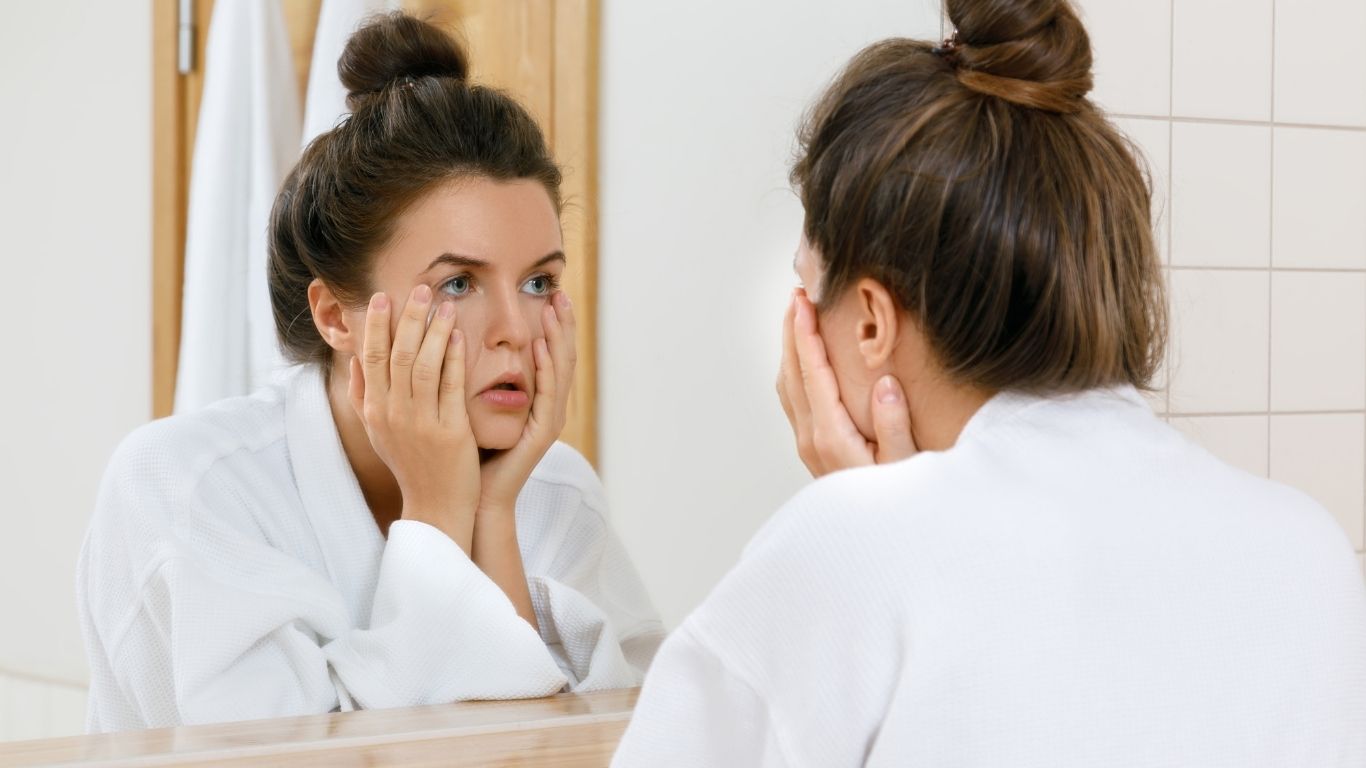 Zmęczona kobieta w białym szlafroku patrzy w lustro i dotyka skóry pod oczami – skutki niedoboru snu na twarzy